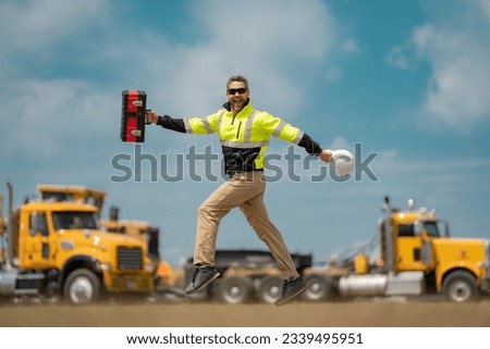 Crazy builder excited jump on site construction. Excited builder construction worker in a safety helmet jumping in front of the trucks. Excited crazy builder man in helmet jump outdoor. Royalty-Free Stock Photo #2339495951