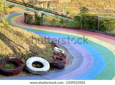 a photography of a rainbow painted track with a roller coaster in the background, there are tires that are sitting on the side of a hill.
