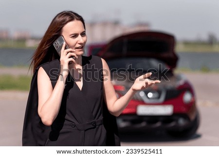 Beautiful girl in a black suit is talking on the phone in front of a red car with an open hood. Calls for technical assistance to fix a car breakdown