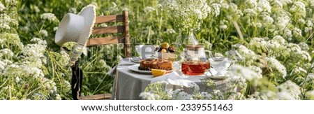 Elegant glamour table setting outdoor in the garden. White porcelain cups, teapot with herbal tea, homemade cherry sweet pie. Family tradition, beautiful place outside. Flower meadow, candles. Banner Royalty-Free Stock Photo #2339551463