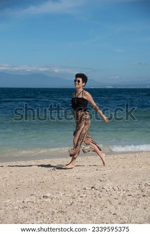 Happy attractive girl running at the beach in summer vacation. Stylish pretty woman with short black hair wears in leopard pant having fun on the tropical beach with copy space.
