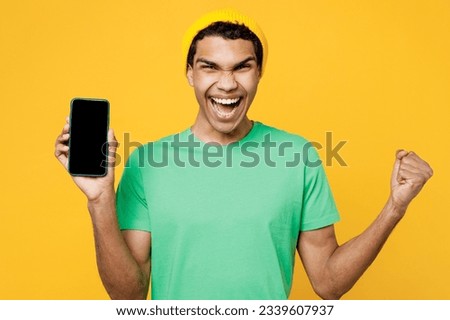 Young man of African American ethnicity wears casual clothes green t-shirt hat hold in hand use mobile cell phone with blank screen workspace area do winner gesture isolated on plain yellow background