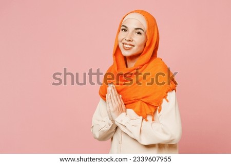 Young arabian asian muslim woman wears orange abaya hijab hold hands folded in prayer gesture isolated on plain light pink background studio portrait. People uae middle eastern islam religious concept Royalty-Free Stock Photo #2339607955