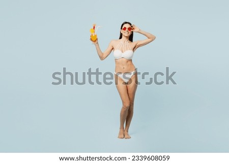 Full body young woman wears swimsuit hold in hand orange juice cocktail show ok okay lower glasses near hotel pool isolated on plain pastel blue background. Summer vacation sea rest sun tan concept