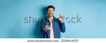 Celebration and holiday concept. Hopeful young man making wish on birthday cake, cross fingers for good luck and smiling positive, standing on blue background.