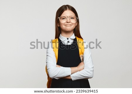 Proud nerdy pupil girl wearing eyeglasses smiling and looking at camera with hands crossed while standing against white background