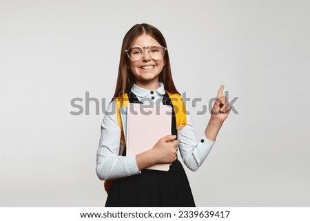 Clever little nerdy girl kid in school uniform and eyeglasses holding books and pointing up, standing isolated over white background
