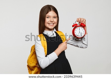 Cheerful nerdy girl in school uniform and yellow backpack holding and pointing at red alarm clock against white background. Don't be late at school.