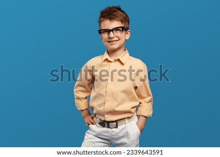 Smart little kid boy wearing nerdy glasses and beige shirt smiling for camera while keeping hands on waist against blue background. Children studio portrait.