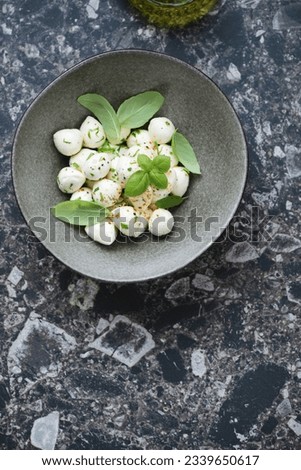 Bowl with marinated mini mozzarella balls and green basil, flat lay on a dark-brown granite background, vertical shot with space
