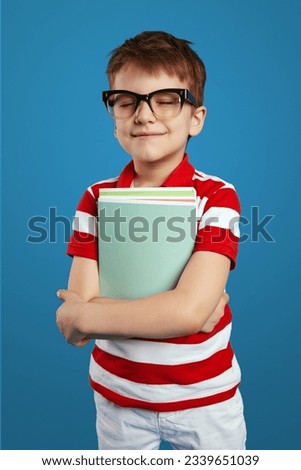 Vertical photo of delighted schoolboy in nerdy eyeglasses smiling and holding textbooks while standing with closed eyes, dreaming about vacation, isolated over blue background.