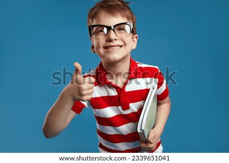 Close up portrait of nerdy boy in retro eyeglasses and red striped shirt smiling and holding textbooks while showing thumb up gesture, standing isolated over blue background