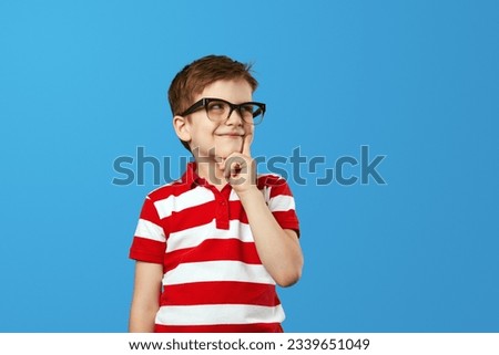 Nerdy boy in eyeglasses and red striped shirt rubbing chin and looking away while thinking near empty space against blue background