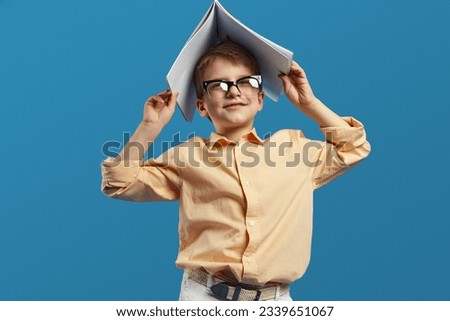 Funny clever schoolboy wearing nerdy eyeglasses and holding open textbook on head while smiling and looking away while against blue background