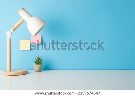 Prepare for classes with this side view picture of a tidy desk featuring desk lamp and tiny flowerpot. Empty blue wall with sticky notes ready for text or promotion Royalty-Free Stock Photo #2339674847