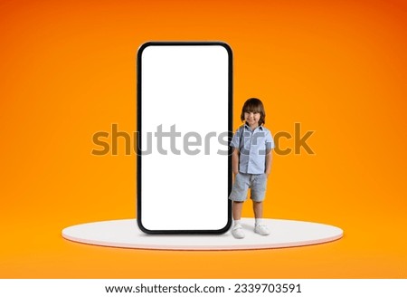 Entertaining mobile app for kids. Cute cheerful little boy pre-school age standing on platform by huge phone with white blank screen, orange studio background, blank space, mockup