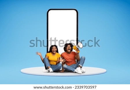 Exciting online offer, deal. Thrilled millennial black couple hipsters posing by huge phone with white blank screen, sitting on platform over blue background, making announcement via loudspeaker
