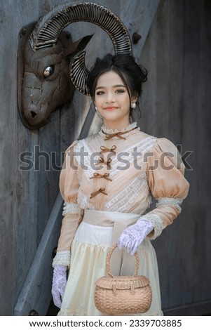 A pretty young Asian teenage girl wearing Victorian or Edwardian-era dresses is happy and looking and smiles at the photographer with a metal goat's head handicraft hanging on the old wooden door.