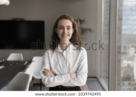 Happy pretty young professional woman posing with arms crossed in modern office boardroom, looking at camera, smiling. Ambitious motivated female business leader heads hot portrait
