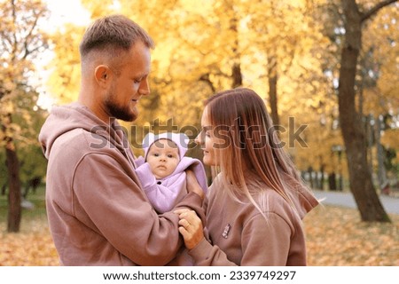 Young mother and father are walking with a newborn baby in the autumn park. Parents in identical tracksuits smile and look at the child. Horizontal photo