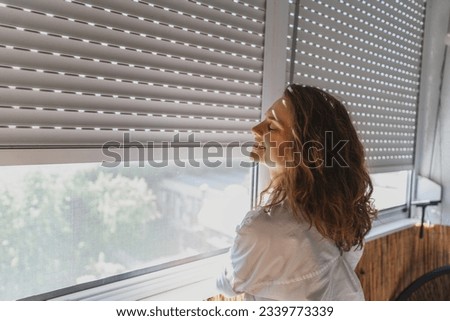 Young woman closing roller shutters on the balcony on a summer day Royalty-Free Stock Photo #2339773339