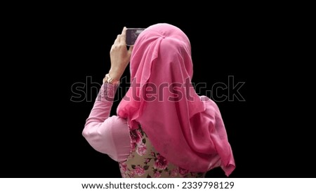 Close up portrait of young Muslim woman back of head wearing pink hijab and dress with red rose motif taking picture and video with phone camera.