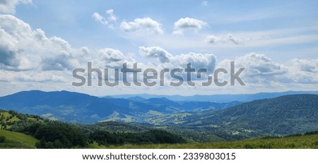Fluffy clouds glide gracefully across the sky, casting playful shadows on the majestic mountains and lush green grasslands below on a sunny summer day. The play of light and shadow creates a mesmerizi