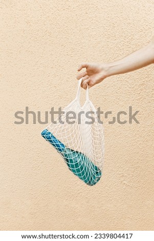 Blue champagne sparkling wine bottle in mesh bag in men hand against light yellow painted wall on street, minimal trend alcohol drink concept, summer stylish lifestyle aesthetic photo, copy space
