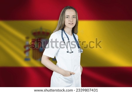 A young female doctor in a white coat and a stethoscope stands on the background of the flag of Spain.