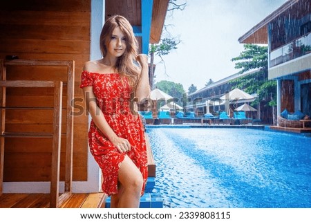 Attractive lady in red dress enjoys poolside relaxation in modern resort. Concept of travel and leisure.
