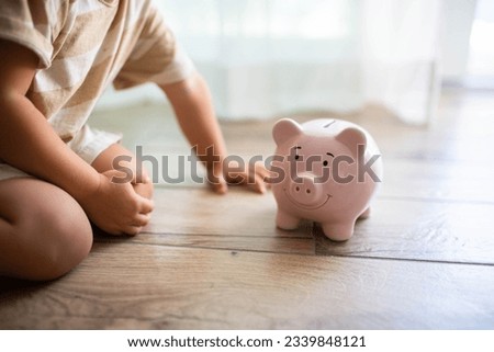Toddler sitting on the floor near a smiling piggy bank. Saving money for kids concept. Royalty-Free Stock Photo #2339848121
