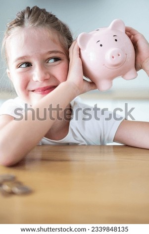 Happy caucasian child girl shaking a smiling pink piggy bank in her hands on a wooden table. Saving money for kids concept. Vertical shot.
