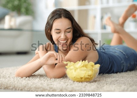 Happy woman laughing and eating potato chips on a carpet at home Royalty-Free Stock Photo #2339848725