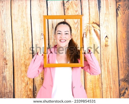 Happy young brunette woman holding an empty picture frame in front of her face against a white background
