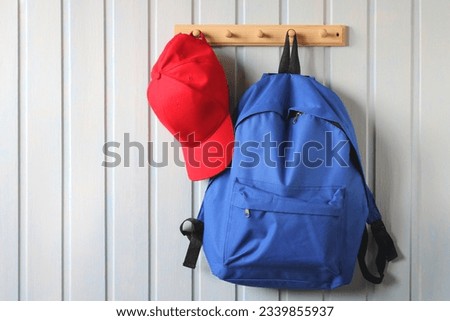 blue backpack and a red baseball cap hang on a wooden hanger on a white board wall.