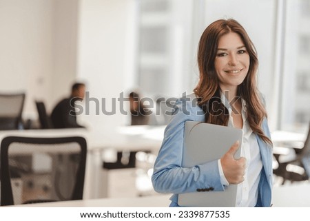 Smiling confident woman wearing stylish business suit holding laptop, looking at camera at modern office. Good looking businesswoman in formal wear posing for picture. Concept of successful business 
