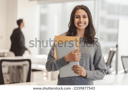 Smiling confident Indian woman wearing casual clothes holding laptop and documents, looking at camera at modern office. Good looking businesswoman posing for picture. Concept of successful business 