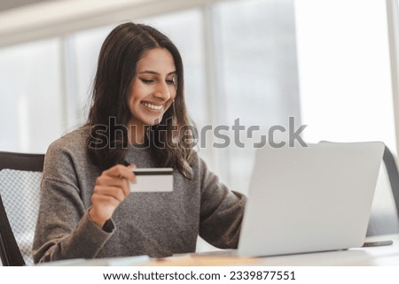 Glad indian woman holding credit card while inputs card number, make online transaction, transferring money at modern office. Happy businesswoman using laptop for e-banking