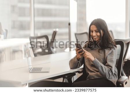 Smiling Indian woman with beautiful hair wearing casual clothes holding mobile phone checking mail in modern office. Attractive female working online, ordering, using mobile app, mobile banking 