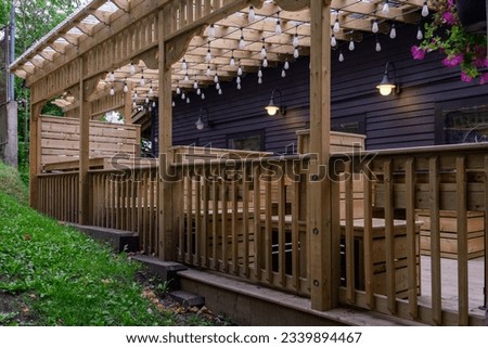 The exterior of a pergola roofed deck of a restaurant. The empty courtyard has wooden table and chairs with stringed patio lights hanging from the rustic pergola made pf pressure treated wood. 