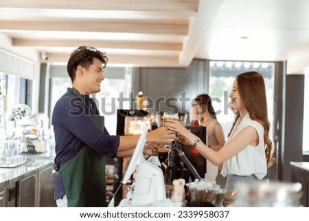 Asian male barista handing iced coffee to female customer over the counter at coffee shop cafe Royalty-Free Stock Photo #2339958037