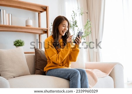 Happy young asian woman relax on comfortable couch at home texting messaging on smartphone, smiling girl use cellphone chatting, browse wireless internet on gadget, shopping online from home Royalty-Free Stock Photo #2339960329