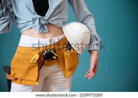 Young female worker with tool belt holding an instrument in hands isolated on background. Pretty caucasian female with tools planning new project. Portrait in studio. Crop view.