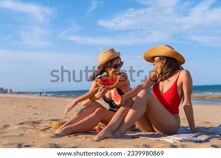 Female friends on vacation at the beach in summer eating a watermelon with the sea in the background, sitting on the sand