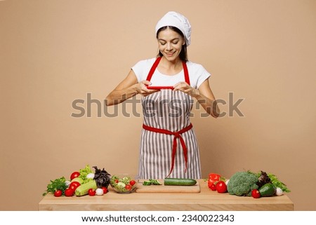 Young housewife housekeeper chef latin woman wear apron toque chefs hat work at table kitchenware taking photo on mobile cell phone isolated on plain pastel light beige background. Process cook food