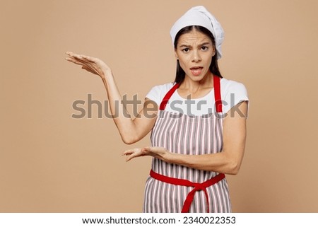 Young indignant angry sad mad housewife housekeeper chef cook baker latin woman wear striped apron toque hat point hand arm aside isolated on plain pastel light beige background. Cooking food concept