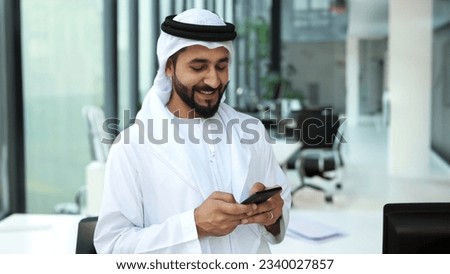 Emirati man inside indoor office wearing Kandura using smart cellphone device. Arab national in disdasha Thobe and Ghutra hair cover cultural Middle Eastern clothing. Royalty-Free Stock Photo #2340027857