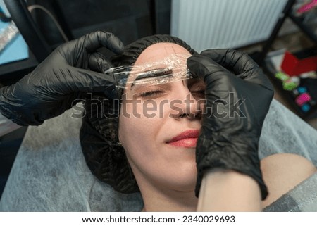 Close-up of the girl's face. An anesthetic cream is applied to the eyebrows. hands of the beautician cover the eyebrows with a transparent film. Royalty-Free Stock Photo #2340029693