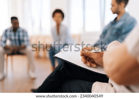 Close-up selective focus shot of unrecognizable mature male psychologist with clipboard having group therapy session with diverse male and female patient sitting in circle. Concept of mental health. Royalty-Free Stock Photo #2340040549