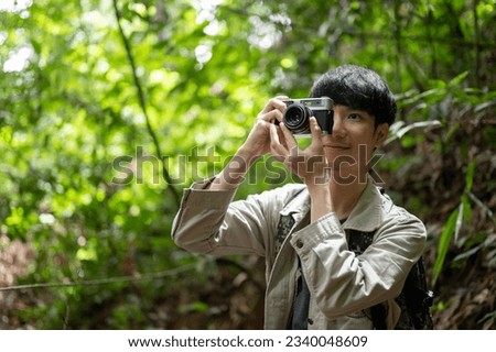 A happy young Asian male hiker takes pictures with his vintage film camera while hiking in the forest alone.